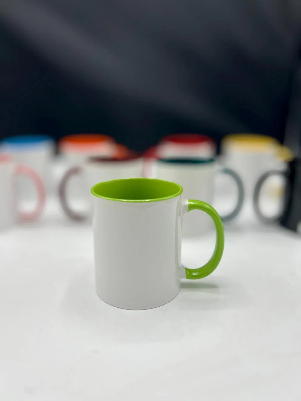 White mug with a green handle on a white surface, with blurred colorful mugs in the background.