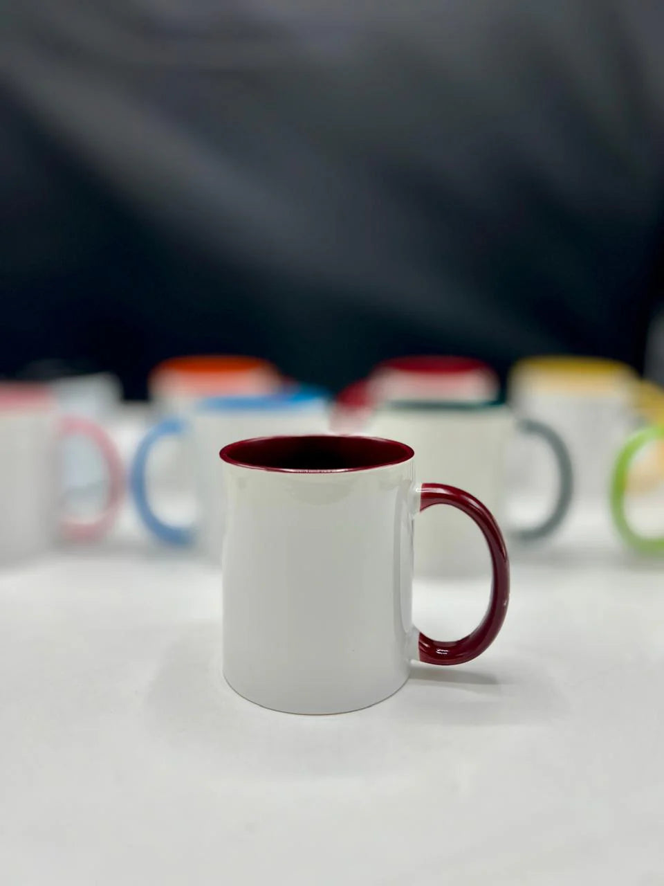 White mug with a red interior and handle on a white surface with blurred colorful mugs in the background.